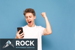 Portrait of a joyful guy in a white T-shirt, rejoices in victory and looks at the screen of a smartphone on a blue background. Joyful guy is using a smartphone and is glad, isolated.